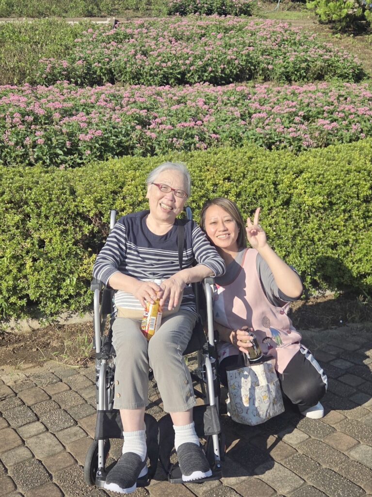 相模原北公園のピンクの花が咲く花壇の前で、スタッフと笑顔で写る車椅子の利用者様
