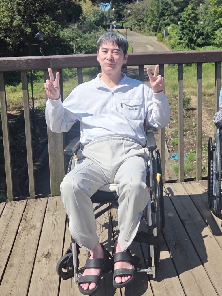 相模原麻溝公園の木のデッキの上で、車椅子に座って両手でピースをする利用者様